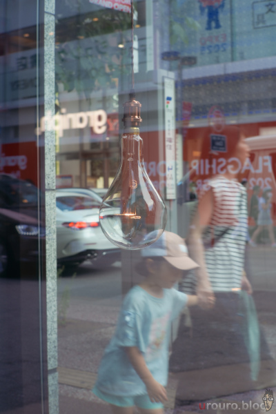 ガラス越しの電球が光っている。奥に通行人も見える。SG-image 35mm F1.2で街スナップ。 フレキシブルカラーはGlimmer。